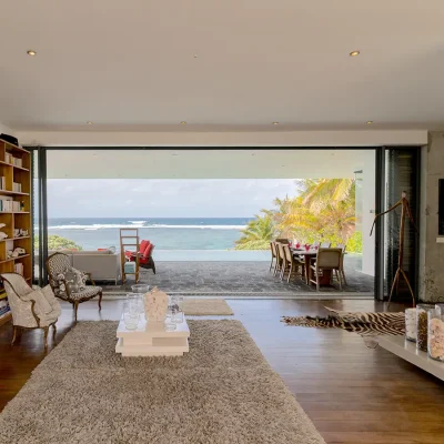 Open plan lounge with a bookcase, two armchairs and a coffee table that opens via stacking doors onto a covered seating area overlooking an infinity pool and ocean blue hues.