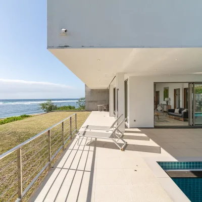 Tiled verandah with two pool loungers overlooking Villa De L'Ocean's rolling lawn and the ocean.