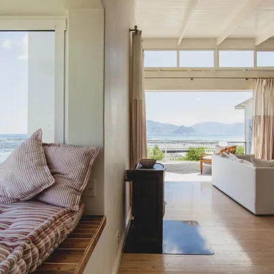 A vast, open-plan living area with a bay window, a free standing fire place and large beige sofa with expansive views of the ocean and coastal mountain range that extends across Pebbles living area through ceiling to floor glass doors.