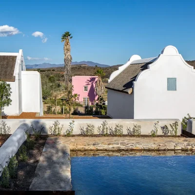 A quaint splash pool protected by a low-rise wrap around whitewashed wall and looking onto whitewashed farm houses with thatched roofs and Buffelsdrift iconic pink wine store with a tall palm tree.