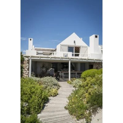 Wooden footpath lined with coastal vegetation leading you to the entrance of Beach House Zonnestraal with whitewashed walls, a thatch roof and a verandah protected by a white bamboo pergola.