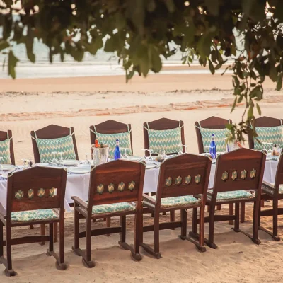 Extensive wooden dining table set with a white linen table cloth and wooden chairs situated on Shela Beach with views of the ocean. The Fort.