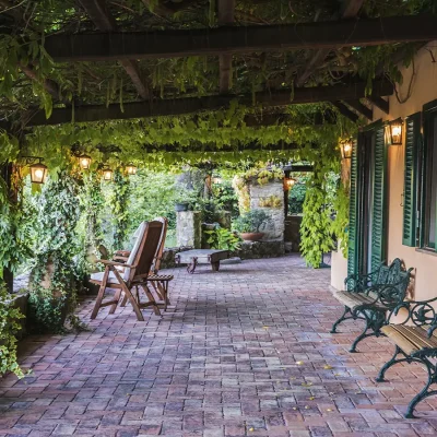 Villa Sunshine's terrace with red brick paving and a pergola wrapped in vines provides a shaded seating area outside the entrance of the tuscan villa that is lined with two benches against the burnt-orange wall and green shutters.