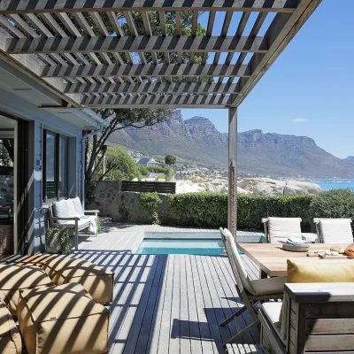 Oasis On Glen wooden deck with yellow sun loungers under a wooden pergola, a built in splash pool and a family sized wooden table with views of Glen beach and the Twelve Apostles mountain range.