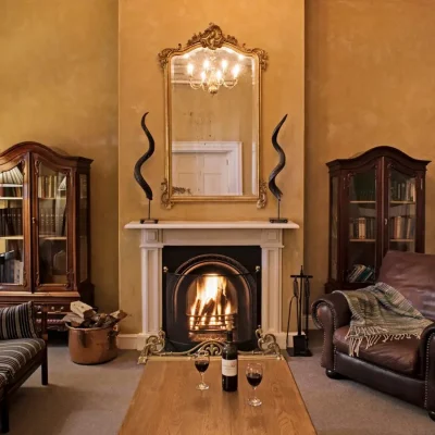 Winter lounge with a central built in fire place with a vintage mirror mounted on beige washed walls reflecting a golden chandelier between two wooden glass cabinets, and surrounded by large leather armchairs and a comfortable striped sofa. Cape Heritage Hotel.