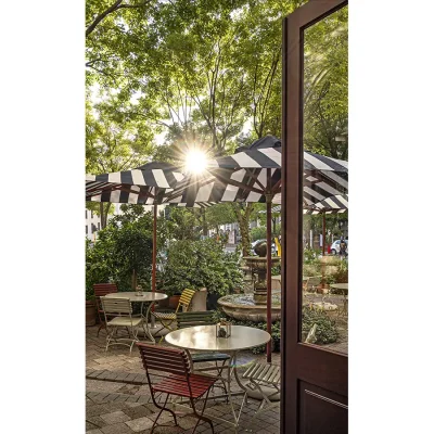 Courtyard with a water fountain surrounded by Parisian Bistro style tables and chairs and black and white striped umbrellas surrounded by luscious green pot plants.