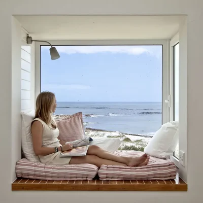 A girl sitting in the bay window with a book in hand and looking out toward the ocean. Pebbles.