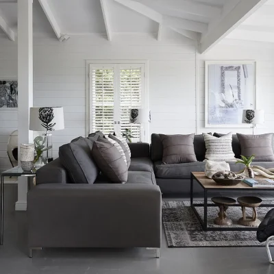 Open plan living area with white cladded walls, large windows, a central free standing fireplace, a soft charcoal 'L-shape' sofa matching the epoxy flooring, and a wooden coffee table with decorative touches. Noordhoek Beach House.