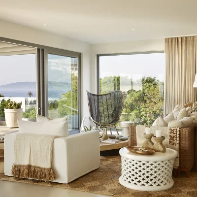 Open plan living area in Walkers Beach Villa furnished with warm beige and brown hues reflecting in the soft beige armchairs and wicker sofa that compliments the woven rug, and decorated with white side tables, a wooden cabinet and wall art that opens onto the patio via large stack back doors with views of the beach and Beacon Isle.