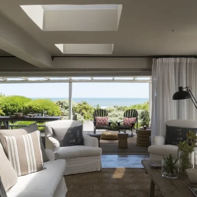 Open plan lounge with soft, white sofa and armchairs warmed by natural light from the skylights and opening onto the deck exposing expansive views of the ocean. Beach House Zonnestraal.