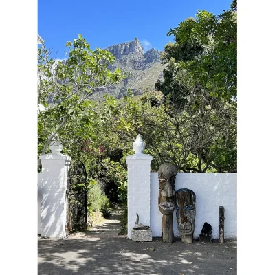 Entrance of Villa Toscana with exposed whitewashed brick walls and wooden statues with a cluster of trees and Table Mountain in the background.