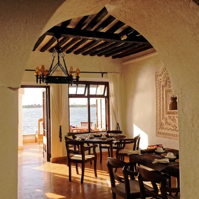 Arched doorway that reveals Peponi Hotel dining area with beige washed walls and wooden beam ceilings, and furnished with round wooden tables and chairs that face views of the ocean.