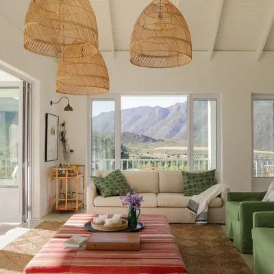 Buchubos living area with a soft beige sofa, green armchairs, a striped ottoman positioned below woven hanging lights and surrounded by large windows and stack doors exposing the mountain range.