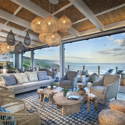 Open plan dining and living area in Lekkerwater Beach Lodge with bamboo ceilings and beige washed walls thaat compliments the beige sofa and armchairs dressed in blue and white scatter pillows, a wooden coffee table and a blue and white floor rug, a large wooden dining table, and large stack back doors that reveals expansive views of the ocean from the deck.