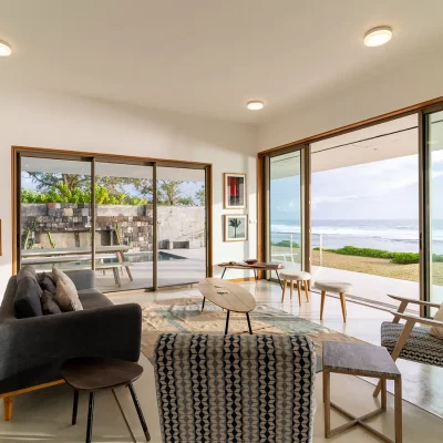 Villa De L'Ocean open plan lounge containing a contemporary couch, two armchairs and an up cycled wooden surfboard coffee table that is surrounded by glass sliding doors and panoramic views of the ocean.