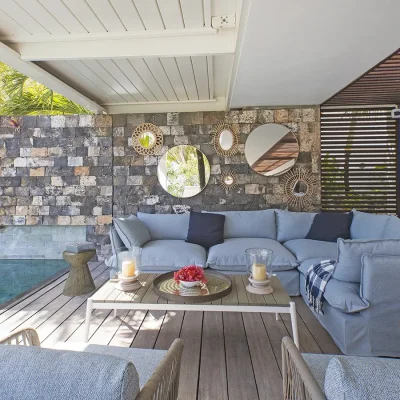 An outdoor corner couch on a covered deck situated next to the pool with a backdrop of a stone wall and decorative wall mirrors.