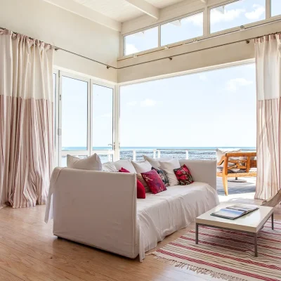 Pebbles living area with pink and white hues that reflect on the white sofa with pink scatter pillows, the striped floor rug and curtains that expose the large glass stack-back doors that expose the wooden deck and views of the ocean.