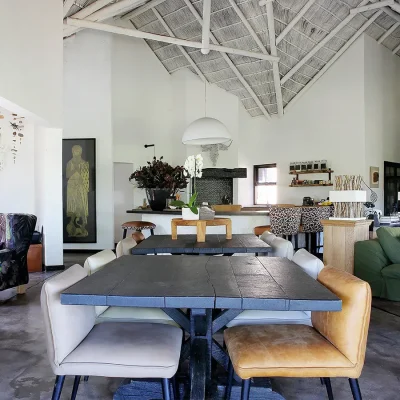 Open plan dining and living area with whitewashed walls and high ceilings, furnished with a central freestanding cast iron fireplace surrounded by wooden dining tables, green sofas and kitchen scattered with varietal decor. Manor House.