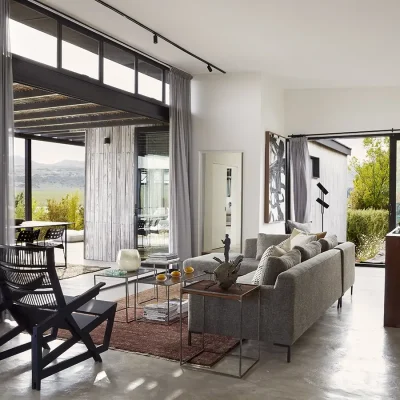 Contemporary open plan living area with white washed walls and soft grey hues reflecting in the soft 'L-shape' sofa and two tiered coffee table that is warmed by a rustic red carpet and the natural light flooding in from the stack-back doors that reveal the grassy plains. The Herenberg.