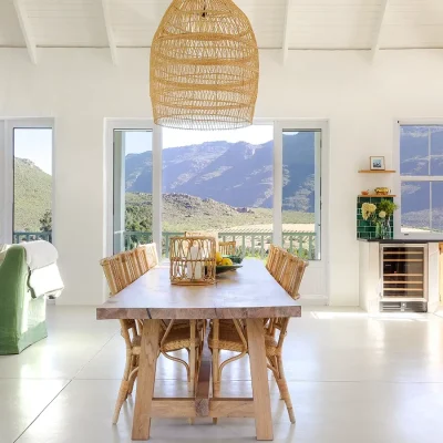 Open plan lounge and dining area with long wooden dining table and wicker chairs facing large glass windows with expansive views of the mountain range.