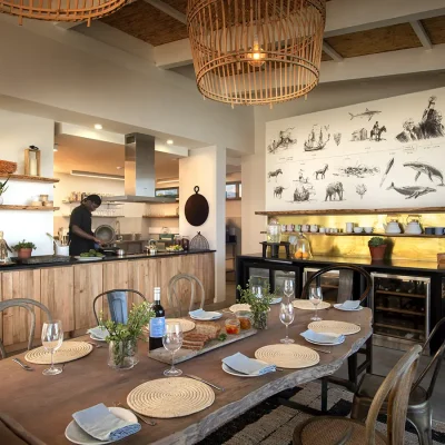 Dining area with a carved wooden dining table, a side drinks cabinet with gold wall paper, and an open plan kitchen with wooden cabinets. Lekkerwater beach lodge.