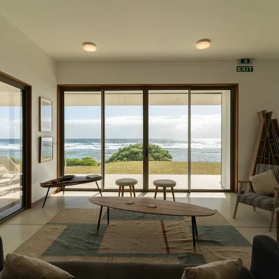 Villa De L'Ocean lounge with upcycled surfboard coffee table and arm chairs with large sliding doors that overlook the rolling lawn and ocean.