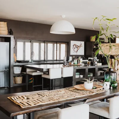 Open planing living and dining space with a carved dark wooden dining table and soft white leather bar stools facing the Blogck kitchen with charcoal walls, a kitchen island with matching leather stools, and stack back windows.