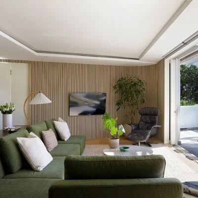 Contemporary lounge with a whitewashed roof and striped wall paper accompanied by a mounted sleek, flat screen television, and furnished with a corner leather armchair, pot plant, a wooden cabinet, and a central green 'L-shape' couch that looks onto Ebb Tide's terrace via large glass sliding doors.