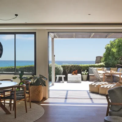 Open plan dining and living area in Oasis on Glen with a round wooden dining table and chairs positioned below a large woven ceiling light and facing the outdoor deck with views of the crystal blue ocean.