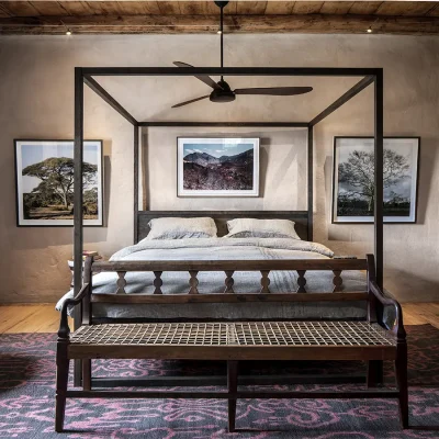 An elegant room in Buffelsdrift with a large wooden four poster bed with grey linen bedding that reflects in the side cabinets, a ceiling fan and original clay walls painted beige.