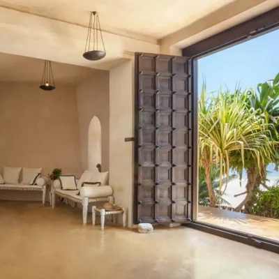 Entrance hallway with beige washed walls that match the polished screed floors and large wooden double doors that opens onto views of the beach and ocean. The Fort.