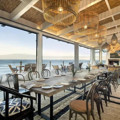 Lengthy carved wooden table in Lekkerwater Beach lodge dining area with a combination of wooden and black bronx dining chairs positioned below large woven hanging lights and facing panoramic sea views and a large fireplace.