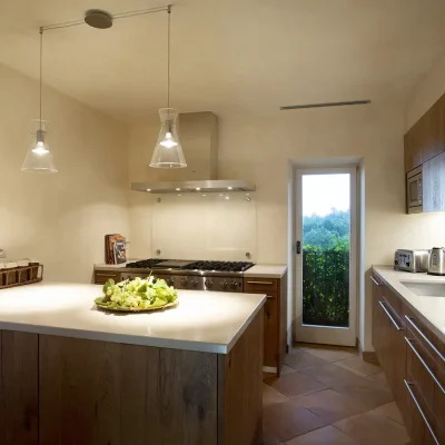 Simplistic kitchen with brown wooden cabinets and spacious, white stone counter tops with a large built-in gas stove and oven, and two hanging ceiling lights that light up the kitchen counter, as well as a glass kitchen door that opens onto Villa Sunshine's back garden.