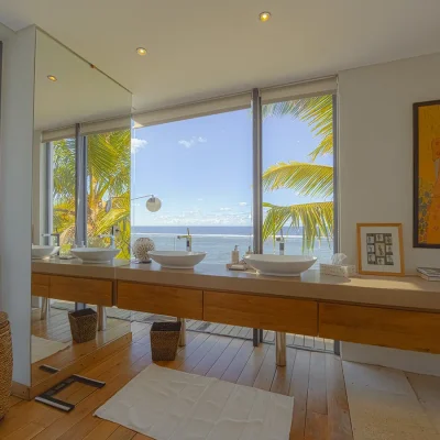 En-suite bathroom with contemporary finishes including wooden floors, double basins, artwork, a door giving access to deck, and a large glass window overlooking the ocean.