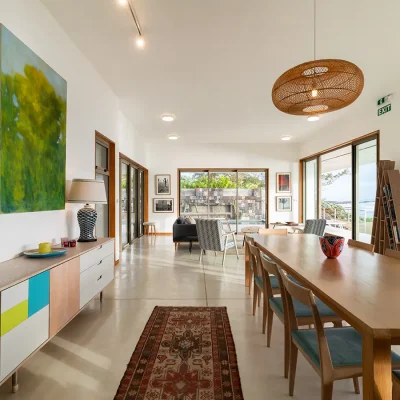 Open plan lounge and dining room with multiple wooden framed entrances, a large wooden dining table, a contemporary bookshelf and cabinet, with ocean views. Villa De L'Ocean.