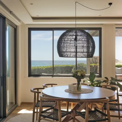 Dining area in Oasis on Glen with a round wooden dining table and chairs positioned below a large woven ceiling light and facing views of the crystal blue ocean from large glass windows.