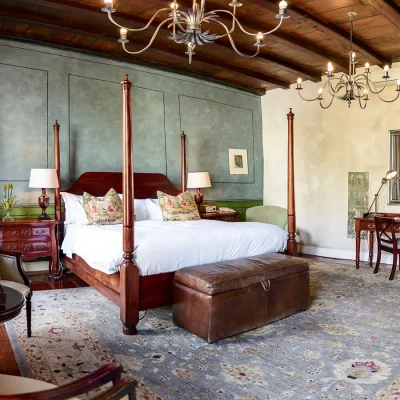 Cape Heritage Hotel Bedroom suite with an antique wooden four poster bed with crisp white linen and floral cushions and complimentary wooden side tables set against a blue washed wall that reflects in the Persian rug.