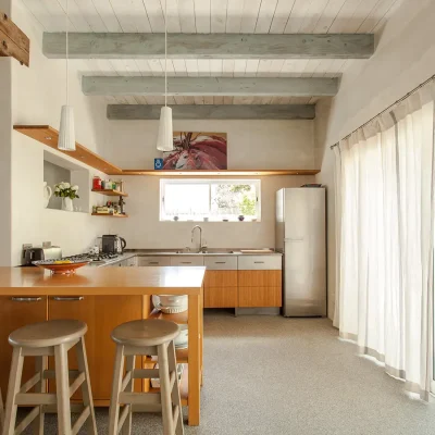 Kitchen with sleek wooden cabinets and counter tops, wrap around shelving with built in lighting and set against beige washed walls with natural light exposure from adjacent glass sliding doors. Pebbles.