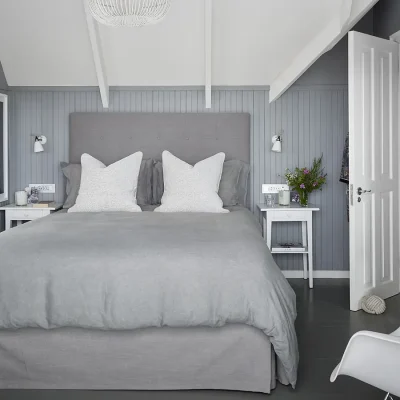 Contemporary bedroom with soft grey and blue hues that reflect on the wall cladding and matches the soft grey headboard and linen, with two white side tables and built in cabinets with mirrors. Noordhoek Beach house.