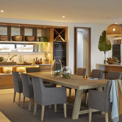 Lengthy wooden dining table with soft brown armchairs surrounded by a wooden glass cabinet and a kitchen island. Walkers Beach Villa.