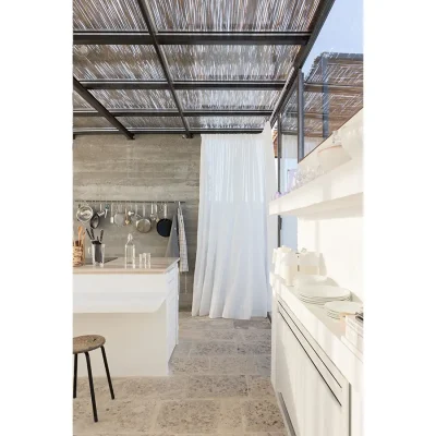 Casa Piana kitchen maintains a serene and minimalistic design with white sleek counter tops, floating shelves and a stone washed wall, cocooned with an expansive glass ceiling shaded by a wooden pergola and wrap around glass doors with linen curtains.