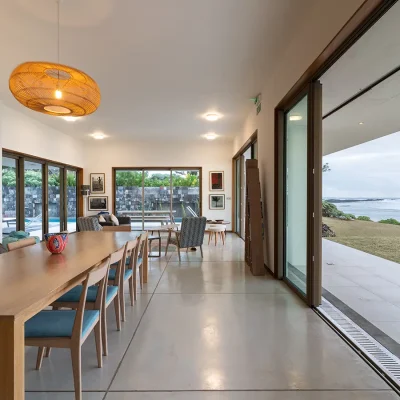 Villa De L'Ocean dining room with sleek wooden table and chairs, and an overhanging woven ceiling light, and large sliding doors opening onto the verandah and garden with ocean view.