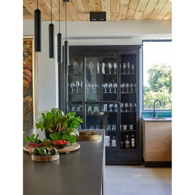 Kitchen with large wooden glasses cabinet, corner window, singular sink and a large island with vegetables.