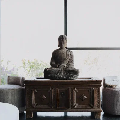 A detailed wooden kist with a buddha statue positioned between two textured off white ottomans. The Blogck.