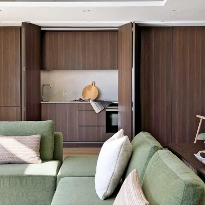 Open plan lounge and dining area in Ebb Tide that is furnished with a green 'L-shape' couch and dining table set, and contains sleek, tall wooden cabinets that stack-back to reveal a contemporary kitchenette.