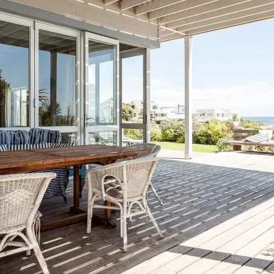 Pebbles wrap around wooden deck with long wooden table surrounded by white whicker chairs and views of the ocean.