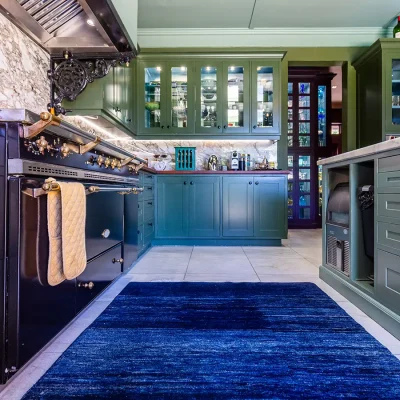 Green kitchen cabinetry with black and gold Victoria cooker and electric blue rug. House Nouveau.