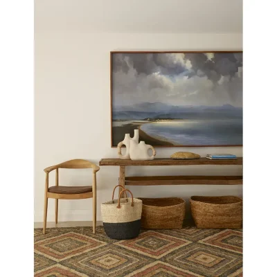 Sleek wooden cabinet that compliments the wooden chair and thin wooden framed painting of Plettenberg Bay coastline positioned in a hallway in Walkers Beach Villa with a warm decorative rug.