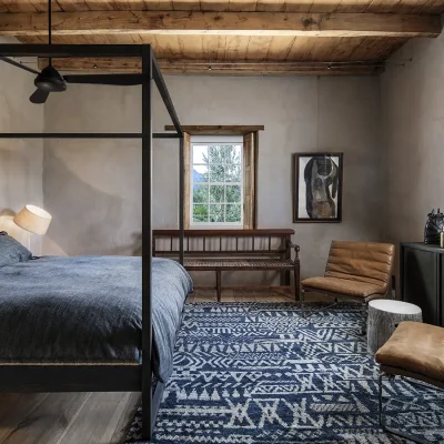 The tasteful but quaint Buffelsdrift room with a four poster bed and charcoal linen bedding that reflects in the opposite cabinet, a ceiling fan, two leather armchairs, large windows with original poplar timber shutters and beige finish.