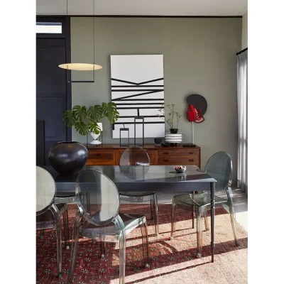 A Gregor Jenkins steel dining table and accompanying Ghost chairs positioned on a rustic red Persian rug that compliments the tone of the dark wood cabinet positioned against a soft olive green wall. The Herenberg.